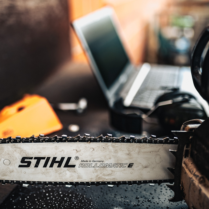 Service für Motorsägenketten schärfen: Eine Kettesäge liegt auf dem Werkstatttisch. Der Fokus liegt auf der Kette, während im Hintergrund ein Laptop zur Fehlersuche zu sehen ist.
